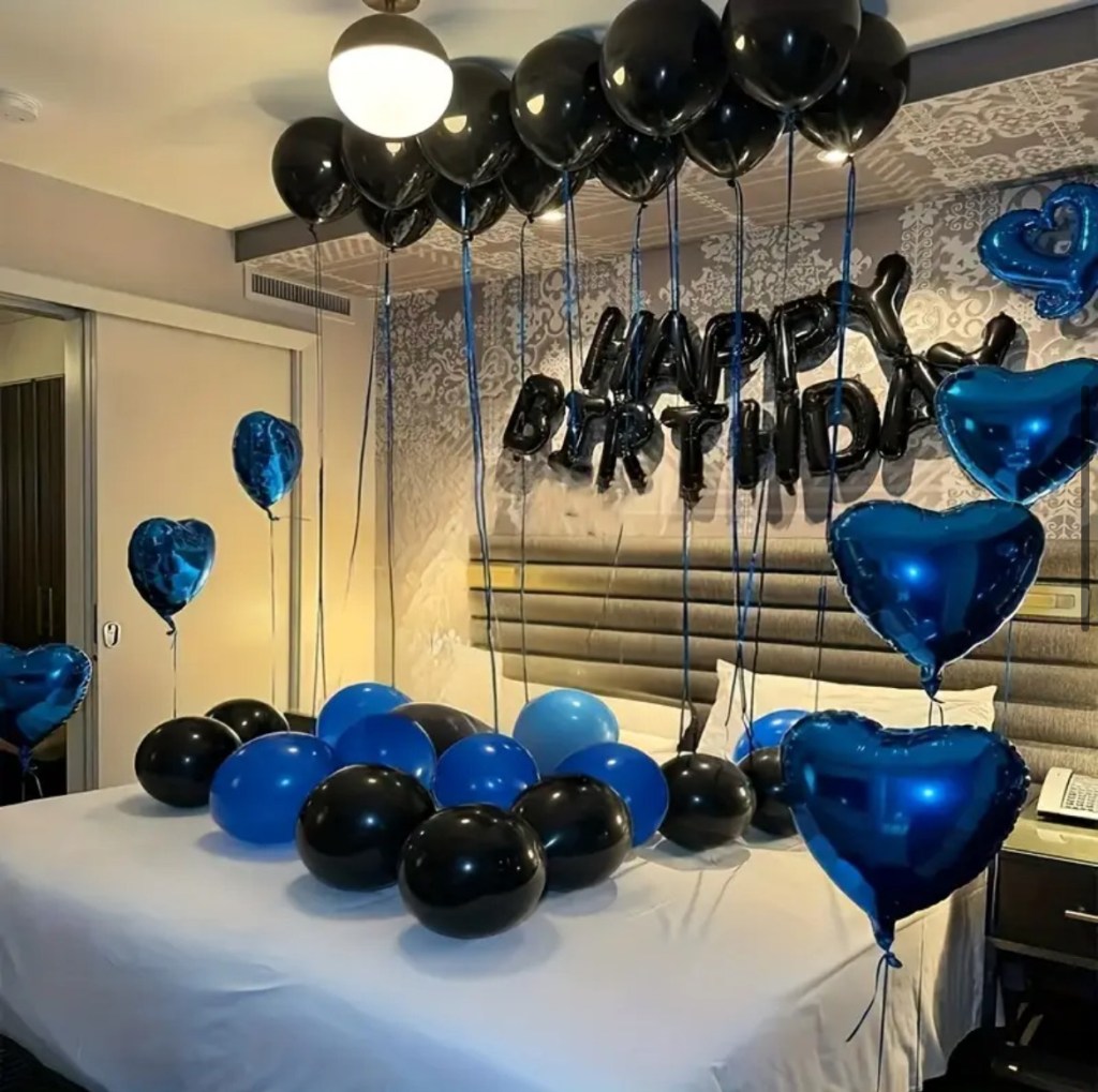Hotel room decorated with black and blue heart shaped and round balloons, with 'Happy Birthday' balloon letters above a bed