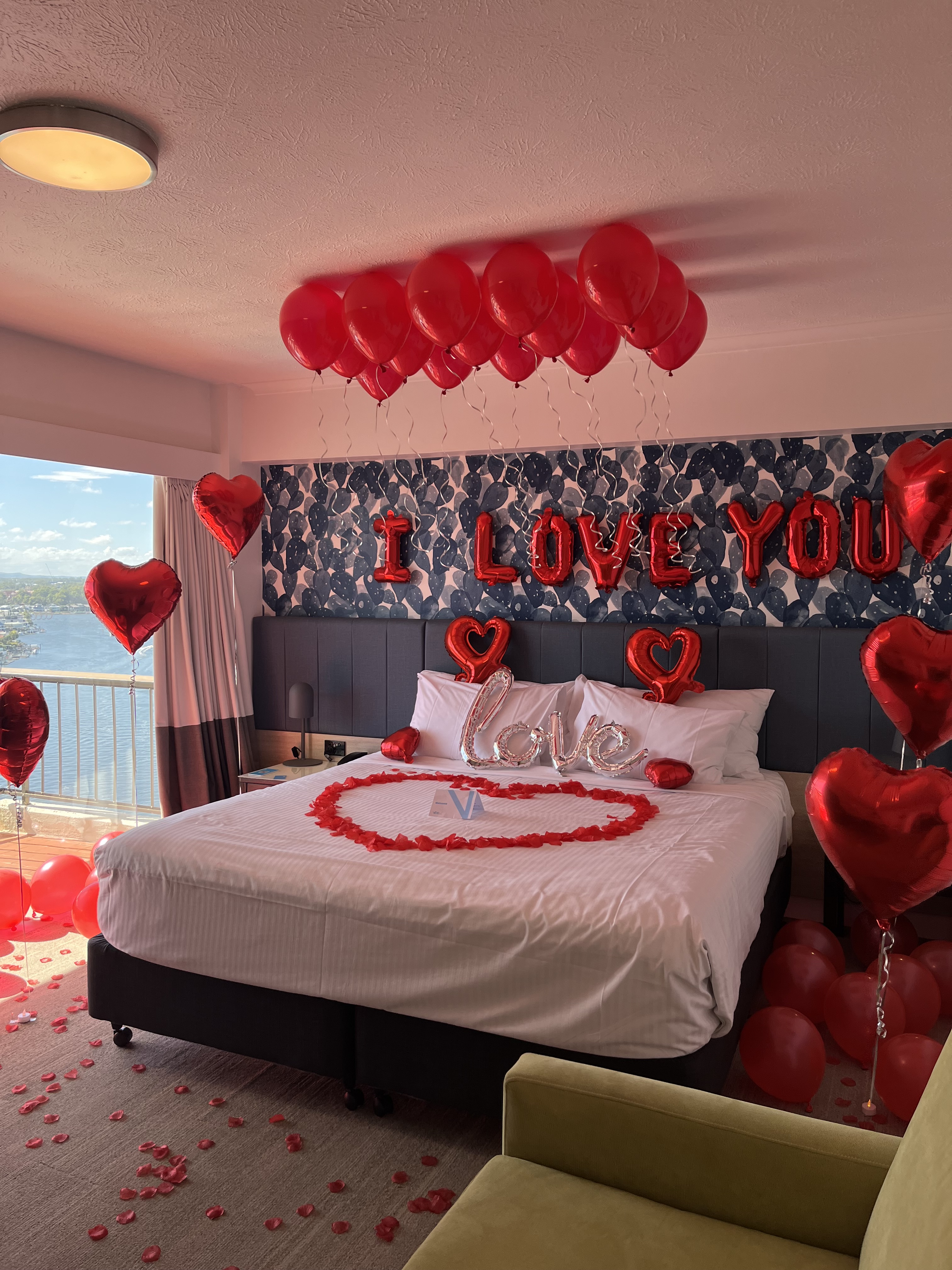 Hotel room with a bed decorated with red heart shaped balloons, rose petals, and 'I LOVE YOU' balloons above.