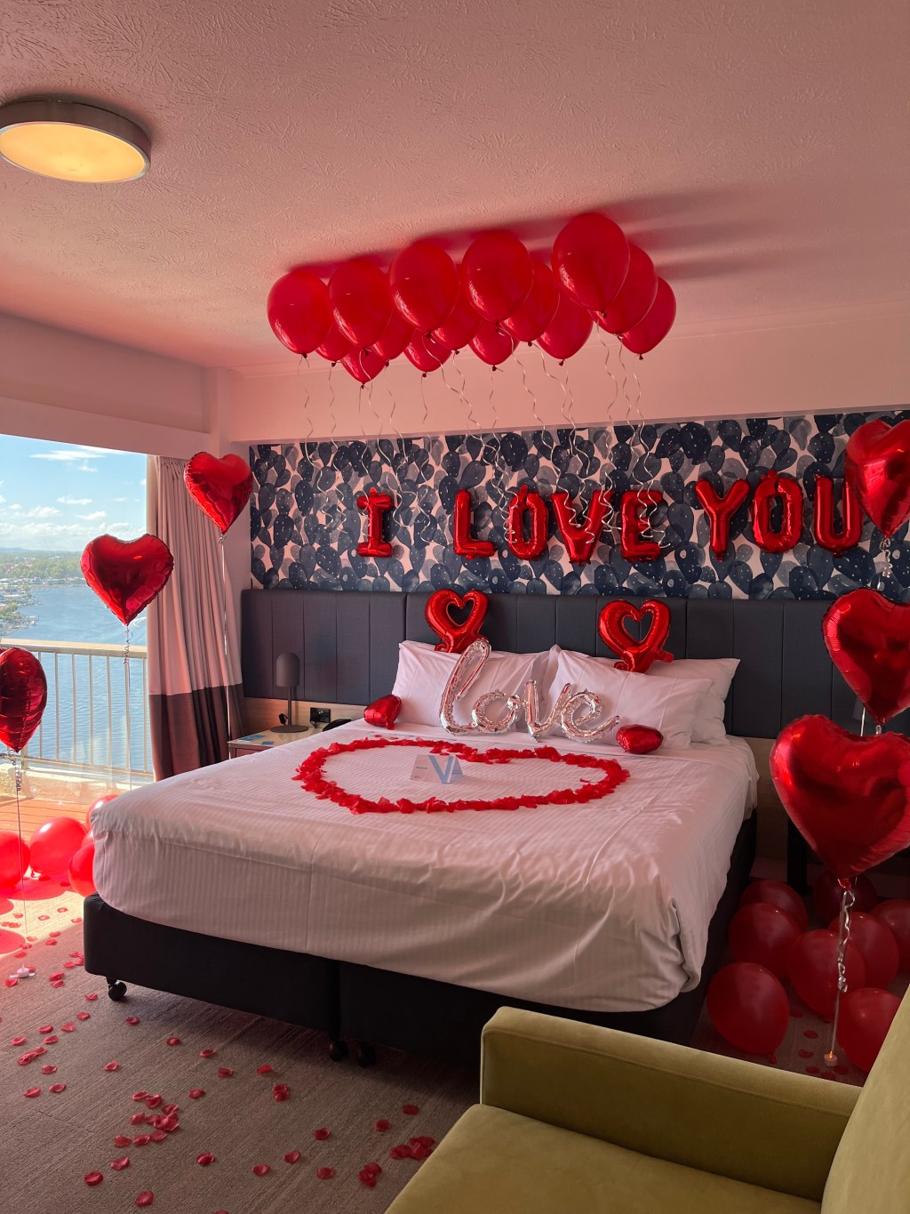 Hotel room with a bed decorated with red heart shaped balloons, rose petals, and 'I LOVE YOU' balloons above.